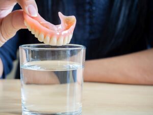 Someone in a dark blue shirt placing dentures into a glass of clear fluid on a wooden table